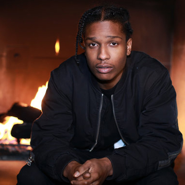 
            Image of rapper A$AP Rocky sitting in front of a fire with his hands clasped together. He is facing the camera and wearing an all black outfit.