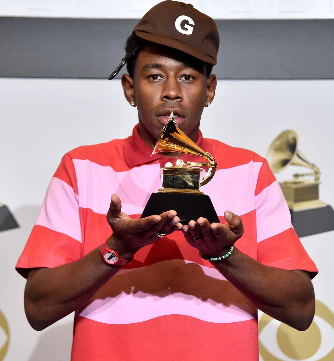 
            Image of rapper and producer Tyler the Creator in a brown hat and striped shirt. He's holding his grammy in the palms of his hand and facing the camera.